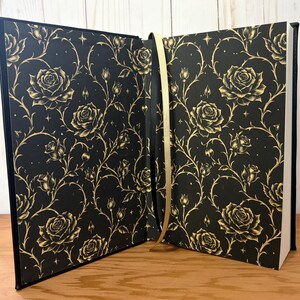 Puede incluir: Libro abierto con una cubierta negra con un patr&oacute;n de rosas y espinas doradas. Las p&aacute;ginas son blancas y se ve una cinta dorada como marcador. El libro est&aacute; abierto sobre una superficie de madera.
