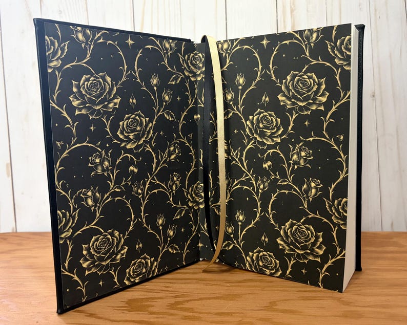 Puede incluir: Libro abierto con una cubierta negra con un patr&oacute;n de rosas y espinas doradas. Las p&aacute;ginas son blancas y se ve una cinta dorada como marcador. El libro est&aacute; abierto sobre una superficie de madera.
