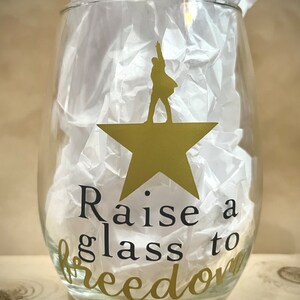K&ouml;nnte beinhalten: Ein klares Weinglas aus Glas mit einem goldenen Stern und einer Silhouette einer Person, die darauf steht. Der Text "Raise a glass to freedom" ist auf das Glas gedruckt.