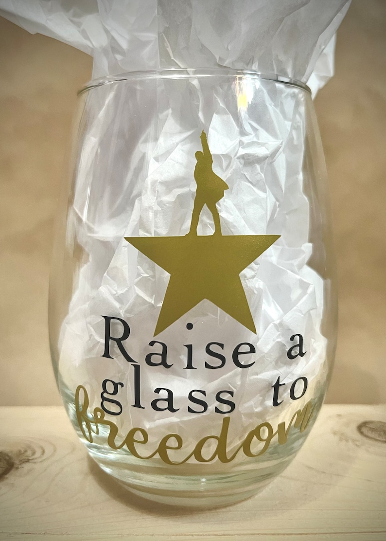 K&ouml;nnte beinhalten: Ein klares Weinglas aus Glas mit einem goldenen Stern und einer Silhouette einer Person, die darauf steht. Der Text "Raise a glass to freedom" ist auf das Glas gedruckt.