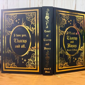 Puede incluir: Libro negro con dise&ntilde;o floral dorado y texto "I love you. Thorns and all." y "A Court of Thorns and Roses" de Sarah J. Maas. El libro tiene un borde dorado y detalles dorados. El libro est&aacute; abierto, mostrando la portada y el lomo.