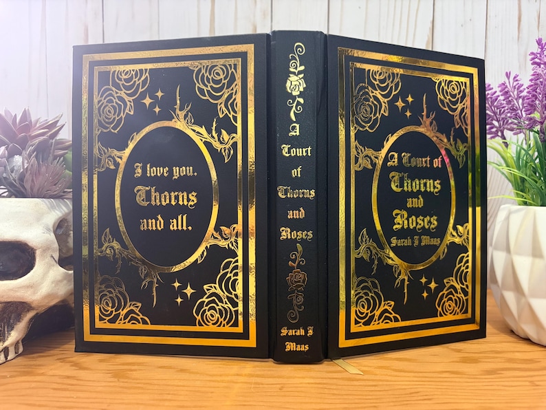 Puede incluir: Libro negro con dise&ntilde;o floral dorado y texto "I love you. Thorns and all." y "A Court of Thorns and Roses" de Sarah J. Maas. El libro tiene un borde dorado y detalles dorados. El libro est&aacute; abierto, mostrando la portada y el lomo.