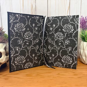 Puede incluir: Diario abierto con una cubierta negra y un patr&oacute;n de rosas blancas. Las p&aacute;ginas est&aacute;n abiertas, revelando el mismo dise&ntilde;o floral. Una cinta blanca como marcador es visible. El diario est&aacute; sobre una superficie de madera, con una calavera y plantas en el fondo.
