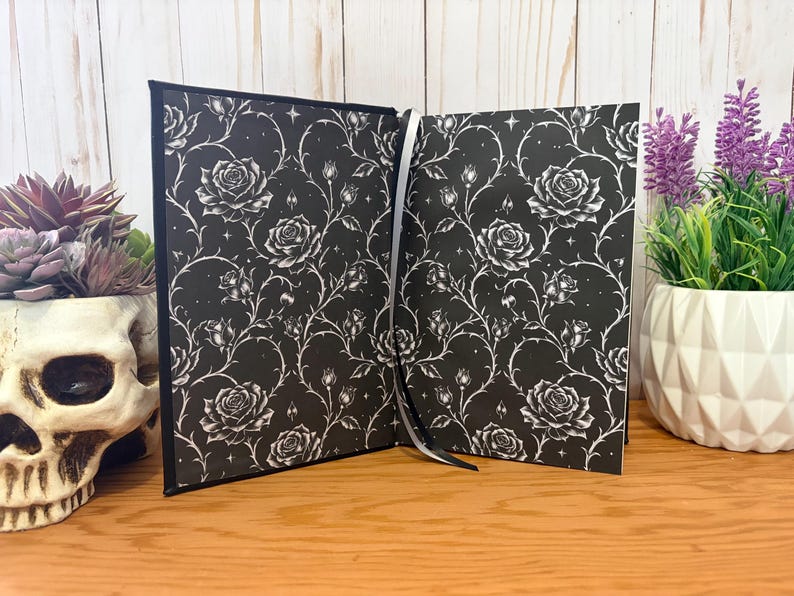 Puede incluir: Diario abierto con una cubierta negra y un patr&oacute;n de rosas blancas. Las p&aacute;ginas est&aacute;n abiertas, revelando el mismo dise&ntilde;o floral. Una cinta blanca como marcador es visible. El diario est&aacute; sobre una superficie de madera, con una calavera y plantas en el fondo.
