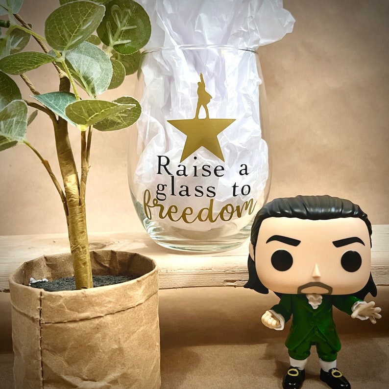 K&ouml;nnte beinhalten: Ein klares Weinglas aus Glas mit einem goldenen Stern und dem Text "Raise a glass to freedom". Im Vordergrund befindet sich eine kleine Figur eines Mannes in einer gr&uuml;nen Jacke und einer schwarzen Hose.
