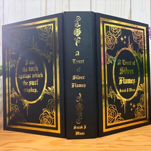 Puede incluir: Un libro negro con detalles dorados. La portada presenta el t&iacute;tulo "A Court of Silver Flames" y el nombre de la autora, Sarah J. Maas. El lomo del libro tambi&eacute;n tiene letras doradas. La portada tiene la cita "I am the rock against which the surf crashes."