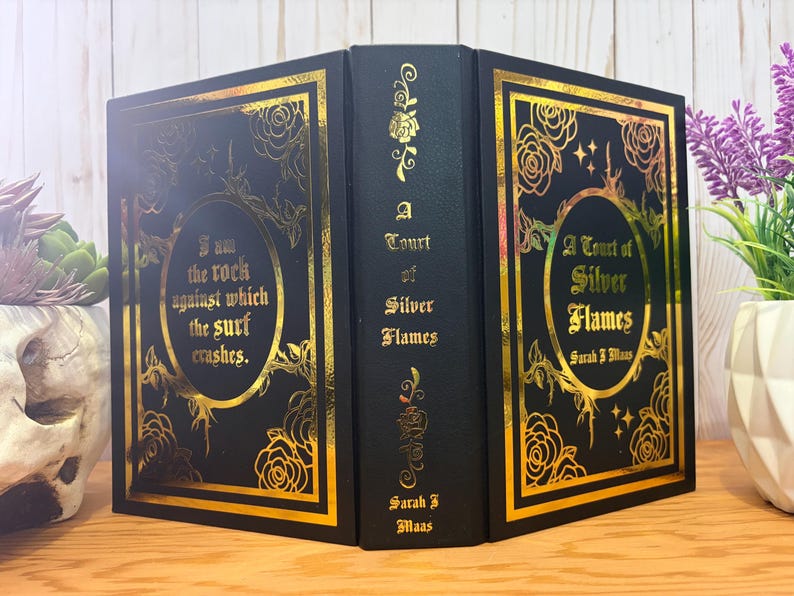 Puede incluir: Un libro negro con detalles dorados. La portada presenta el t&iacute;tulo "A Court of Silver Flames" y el nombre de la autora, Sarah J. Maas. El lomo del libro tambi&eacute;n tiene letras doradas. La portada tiene la cita "I am the rock against which the surf crashes."