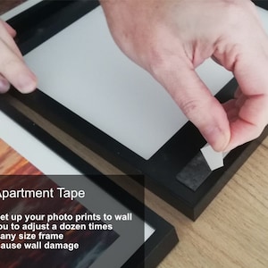 Op de afbeelding: Een hand houdt een stuk doorzichtig plakband over een zwart fotolijst. De tekst "Apartment Tape" is zichtbaar onder de lijst. De tekst onder de titel luidt: "Eenvoudig om uw fotoprints aan de muur te bevestigen. Kan een dozijn keer worden aangepast. Knipbaar om te passen op elke lijstmaat. Veroorzaakt geen schade aan de muur."