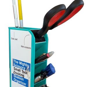 May include: A teal and white tool organizer with various tools. The organizer has compartments for scissors, pens, and other small tools. The text on the organizer reads "The Mighty Scissors Small Tool Docking Station Organizer."