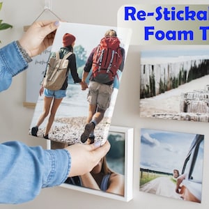 May include: A person is using re-stickable foam tape to hang a framed photo on a wall. The photo shows a couple walking on a beach. There are other framed photos on the wall.