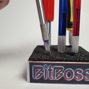 May include: A set of five utility knives with different colored handles, including red, blue, and yellow, are displayed in a black foam holder with the text "BitBOSS" printed on the front.