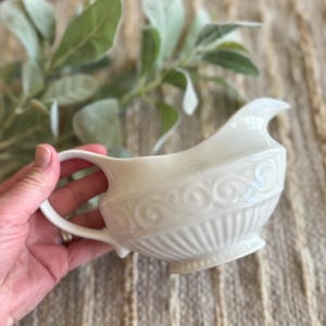 Op de afbeelding: Witte gravyboot van keramiek met een decoratief swirl-ontwerp. De boot heeft een brede, ronde basis en een tuit aan de ene kant.