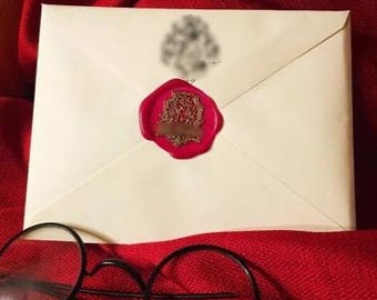 Könnte beinhalten: Ein cremefarbener Umschlag, versiegelt mit einem roten Wachssiegel. Der Stempel hat ein detailliertes Design und ein braunes rechteckiges Etikett. Eine runde, schwarz gerahmte Brille befindet sich im Vordergrund. Der Hintergrund ist ein tiefroter Stoff.