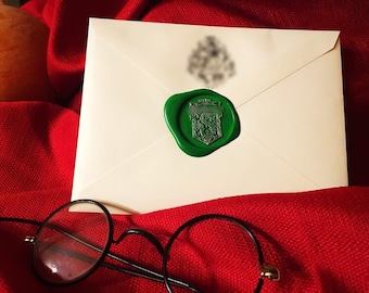 May include: A cream-colored envelope sealed with a green wax stamp featuring a crest design. The envelope rests on a red fabric, with a pair of round, black-framed glasses in the foreground. A candle is partially visible in the background.