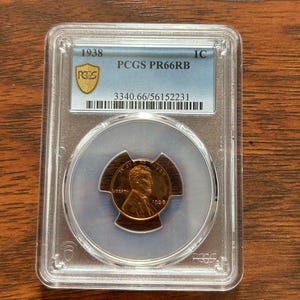 May include: A 1938 Lincoln cent coin, graded PCGS PR66RB, encased in a clear protective holder. The coin features a profile of Abraham Lincoln and the year 1938. The holder has a blue label with the PCGS logo and serial number.