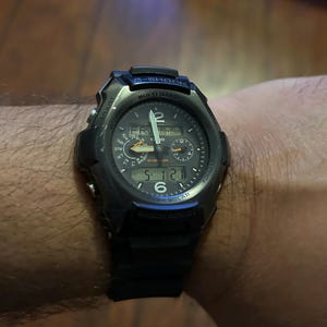 Może przedstawiać: Czarny cyfrowy zegarek G-Shock z okrągłą tarczą, z wieloma tarczami i cyfrowym wyświetlaczem pokazującym godzinę. Zegarek ma czarny pasek i jest noszony na nadgarstku. Zegarek ma napis "MULTI BAND" i "SOLAR".