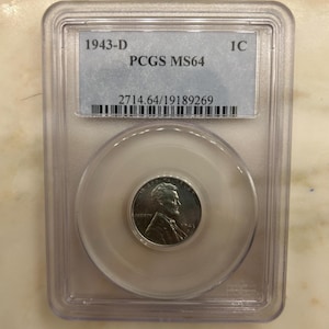 May include: A 1943-D Lincoln cent coin, encased in a clear plastic holder. The coin shows a profile of Abraham Lincoln and the word "LIBERTY". The holder has a label with "1943-D", "PCGS MS64", and a barcode.