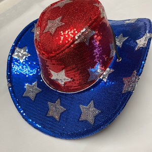 May include: A cowboy hat covered in red, white, and blue sequins. The crown is red with silver sequined stars, while the wide brim is blue with silver sequined stars. A thin black chin cord is attached to the side of the hat crown.