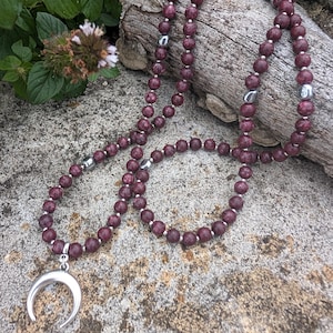 Può includere: Un set di collana e bracciale con perline bordeaux con dettagli argentati. La collana presenta un ciondolo a forma di mezzaluna. I gioielli sono esposti su una superficie di pietra con un tronco di legno sullo sfondo.