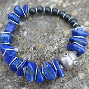 Peut inclure: Bracelet composé de pierres de lapis-lazuli bleu, de perles noires et d'éléments métalliques argentés. Un perle sphérique argentée est également incluse. Le bracelet est présenté sur une surface texturée de tons neutres.