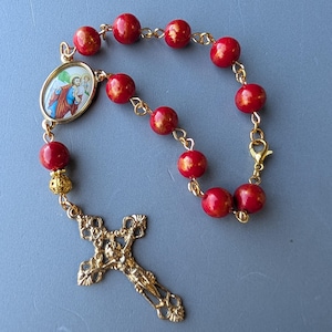 May include: A rosary featuring red beads with gold detailing, linked by gold-coloured chains. It includes a gold-coloured cross and a small oval pendant with an image. The beads have a speckled gold design.