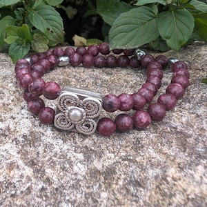 May include: A double-strand bracelet with deep burgundy beads. The bracelet has a square, silver-toned clasp with an intricate floral design. The beads are approximately 0.8 cm in diameter.