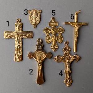 Puede incluir: Una colección de seis colgantes religiosos dorados, que incluyen crucifijos y un dije de la Virgen María. Los colgantes varían en diseño, con detalles intrincados y tamaños variables. El fondo es gris liso.