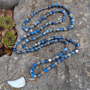 Può includere: Una lunga collana con perline blu e grigie maculate, intervallate da perline di metallo color argento. La collana ha un ciondolo bianco curvo. La collana è esposta su una superficie di pietra grigia.