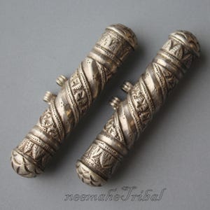 Könnte beinhalten: Zwei silberne, kunstvoll gravierte, zylindrische Behälter mit einem gedrehten Design. Die Behälter haben eine abgerundete Oberseite und eine kleine Schlaufe an der Unterseite.