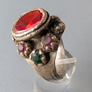 Puede incluir: Anillo de plata con una gran gema roja ovalada y varias gemas más pequeñas rojas y verdes. El anillo tiene una apariencia envejecida y antigua.