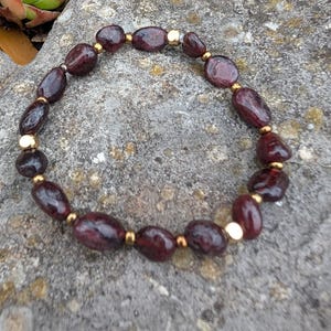 May include: A bracelet made of dark red, oval-shaped beads and small gold-coloured beads. The bracelet is displayed on a textured, grey stone surface. The jewellery piece showcases a natural colour and design. The bracelet is a piece of jewellery.