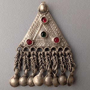 Peut inclure: Un pendentif triangulaire en métal argenté avec une surface texturée, orné de trois pierres rouges circulaires et d'une pierre verte centrale. La base du pendentif comporte huit chaînes pendantes, chacune terminée par une petite breloque ronde.