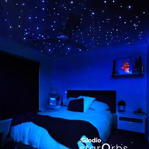 May include: A bedroom with a blue ceiling covered in white fiber optic stars. The bed has a blue blanket and white pillows. The Glodio starOrbs logo is visible in the bottom right corner of the image.