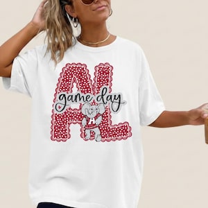 May include: A white short-sleeve t-shirt featuring a large red graphic with a white polka dot pattern forming the letters AL. The text game day is written in black script across the center, accompanied by a cartoon elephant wearing a red jersey.