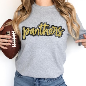 Może przedstawiać: Szara bluza z napisem "Panthers" w złotych literach z brokatem. Bluzę trzyma osoba, która trzyma również brązowy futbol.