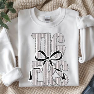 Puede incluir: Una sudadera blanca de cuello redondo con la palabra TIGERS en letras grandes de estilo bloque con un patrón de rayas verticales grises y blancas. Un lazo gráfico negro está centrado sobre el texto, delineado con un borde blanco festoneado.