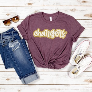 May include: A maroon t-shirt with the word "chargers" in gold glitter lettering. The shirt is paired with blue jeans, white sneakers, and brown sunglasses.