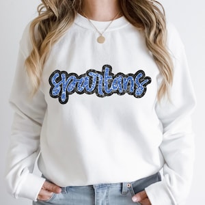 Puede incluir: Sudadera blanca con un gráfico de 'Spartans' en purpurina azul y negra.