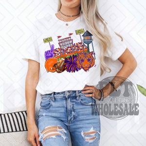 May include: White t-shirt with a vibrant graphic of a football stadium, a tiger, a helmet, and a pom-pom. The design uses orange, purple, and white, ideal for sports enthusiasts.