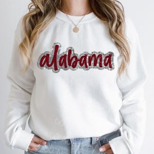 Könnte beinhalten: Weißer Pullover mit einem roten Glitzerdesign, auf dem "alabama" steht.