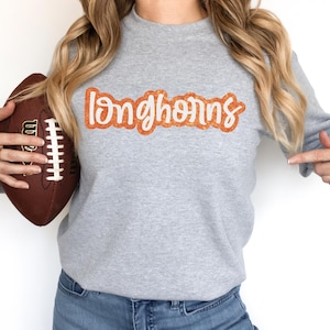 Könnte beinhalten: Ein graues Sweatshirt mit dem Wort "Longhorns" in Orange auf der Vorderseite. Eine Person hält einen braunen Football.