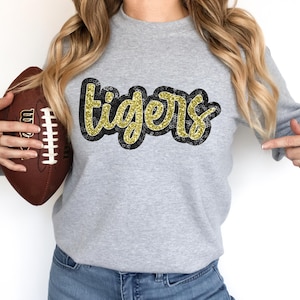 May include: A gray sweatshirt with the word "tigers" in gold glitter letters on the front. The sweatshirt is being held by a person who is also holding a brown football.