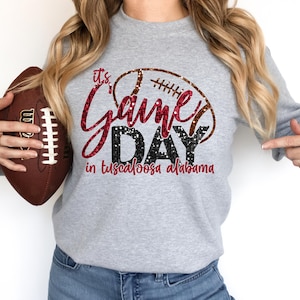 Könnte beinhalten: Ein graues Sweatshirt mit einem roten und schwarzen Glitzerdesign, auf dem "It's Game Day in Tuscaloosa Alabama" steht. Ein Football ist im Bild.