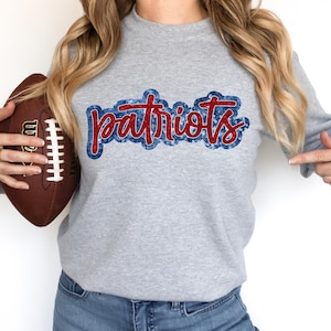 May include: A grey sweatshirt with a red and blue "Patriots" graphic. The sweatshirt is being held by a person who is also holding a brown football.