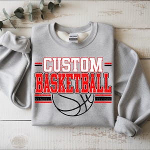 May include: A light grey crewneck sweatshirt featuring the red and white text CUSTOM BASKETBALL above a black line drawing of a basketball. The design includes horizontal red lines and a Gildan brand label at the inner neckline of the garment.
