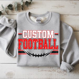 Op de afbeelding: Een lichtgrijs sweatshirt met ronde hals en de in rood en zwart gedrukte tekst CUSTOM FOOTBALL op de voorzijde. Onder de tekst staat een zwarte afbeelding van veters van een football. Het kledingstuk heeft lange mouwen en een Gildan-label.
