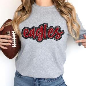 May include: A gray sweatshirt with a red and black "Eagles" design. The sweatshirt is being held by a person who is also holding a brown football.
