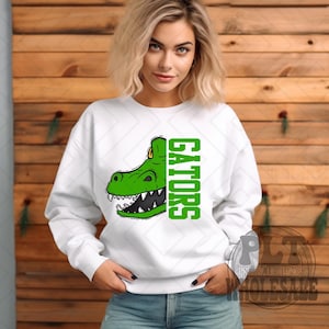 May include: White sweatshirt with a green alligator head graphic and the word "GATORS" in green vertical text. The alligator has sharp teeth. Set against a wooden background.