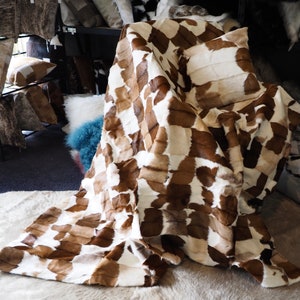 May include: A brown and white patchwork fur throw blanket made of cowhide. The blanket is folded and draped over a chair.