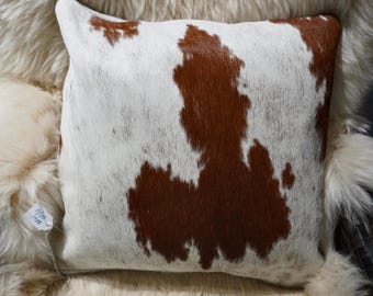 Puede incluir: Una almohada de piel de vaca blanca y marrón con una mancha marrón en el centro. La almohada está sentada sobre una manta de piel blanca.